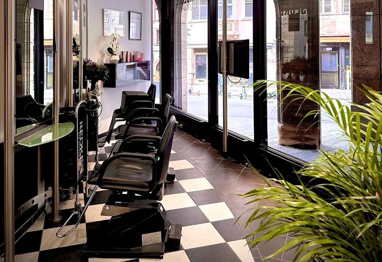 Friseursalon Adler & Herz mit schwarz-weißem Schachbrettboden, schwarzen Frisierstühlen an der Spiegelwand und Fensterfront zur Straße